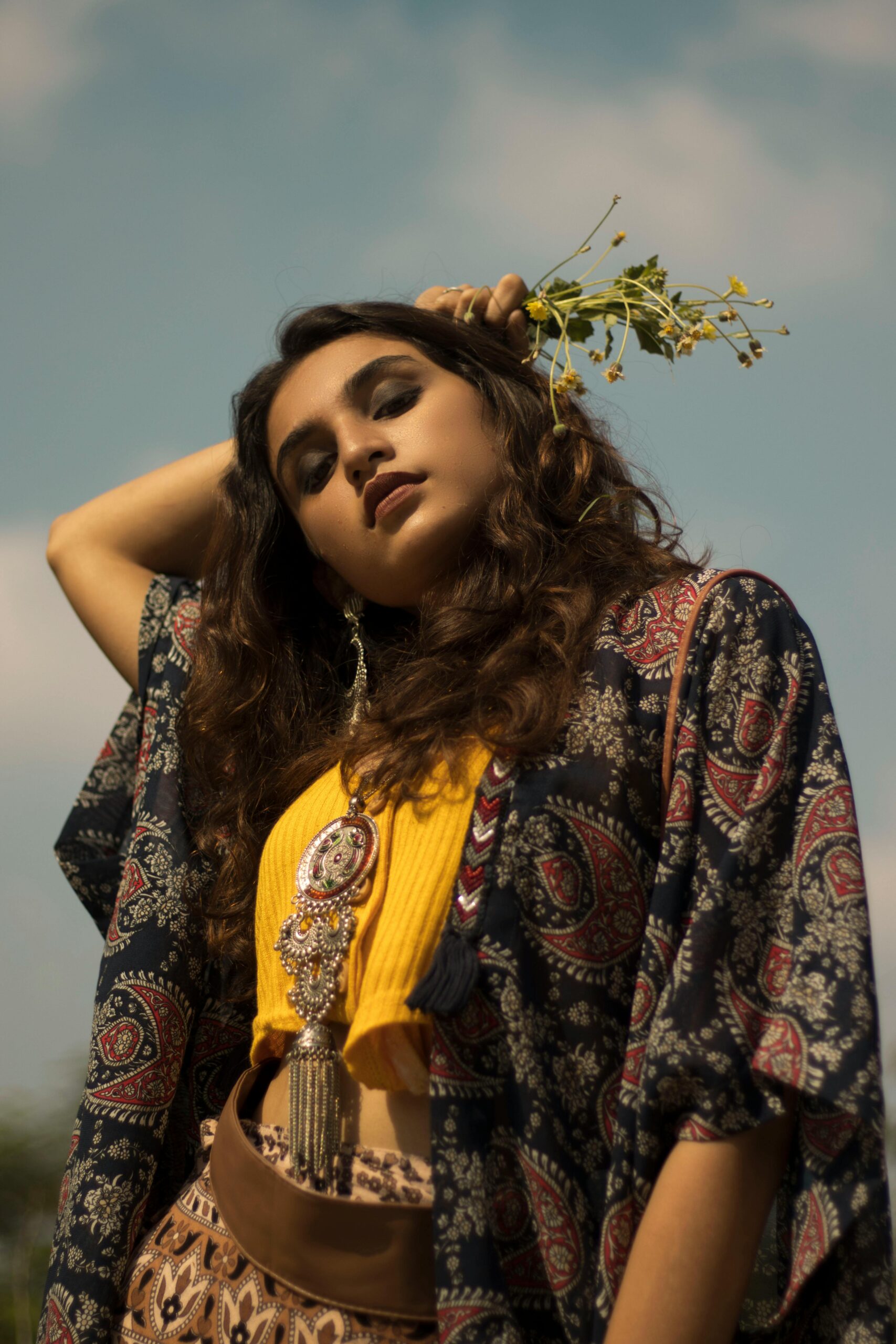 A fashionable South Asian woman with flowers poses stylishly under a blue sky.